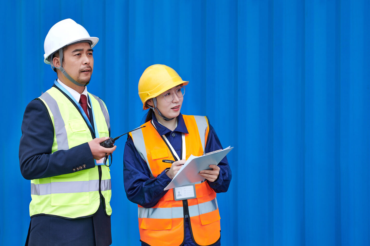 Two warehouse workers wearing hardhats and safety vests. One worker is writing while the other is observing.