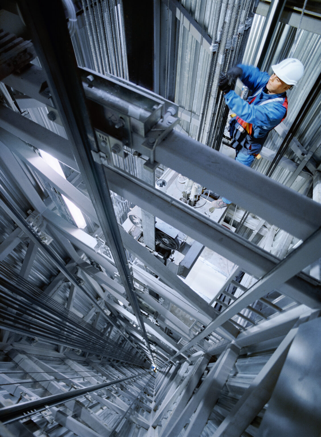 Worker wearing a white hardhat and high-visibility vest, standing inside a newly constructed building. The worker is observing or inspecting the construction site, with exposed beams and unfinished walls visible in the background, indicating an active construction phase