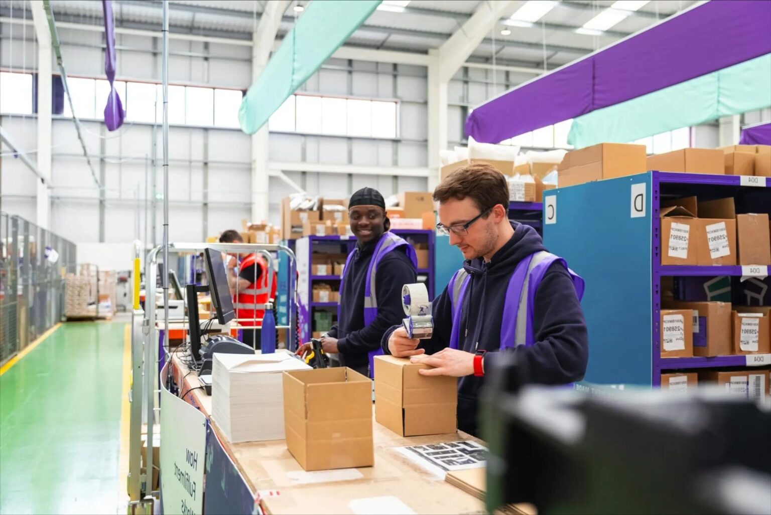 Warehouse workers standing in a line, efficiently packing boxes on a production line. Each worker is focused on their task, with some sealing boxes and others preparing items for shipment. The environment is organized, with shelves of inventory and packing materials visible in the background.
